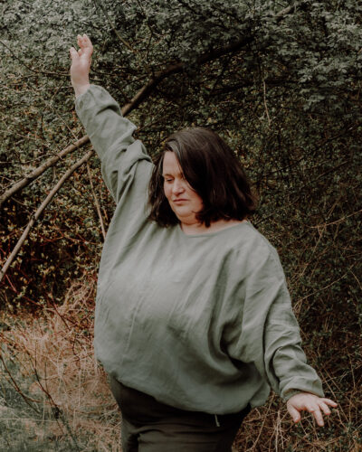 Lniana bluzka damska Comfy w kolorze zgaszonej zieleni plus size w dynamicznym ujęciu w plenerze.